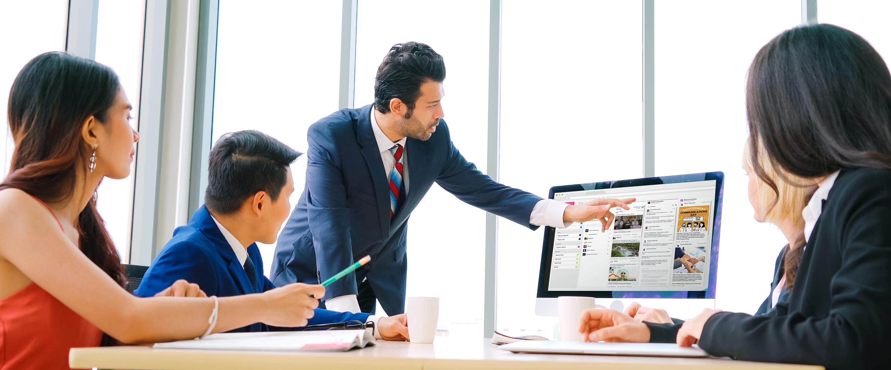 A group of professionals in a meeting room with a man in a suit pointing at a computer screen displaying social media management software. SND